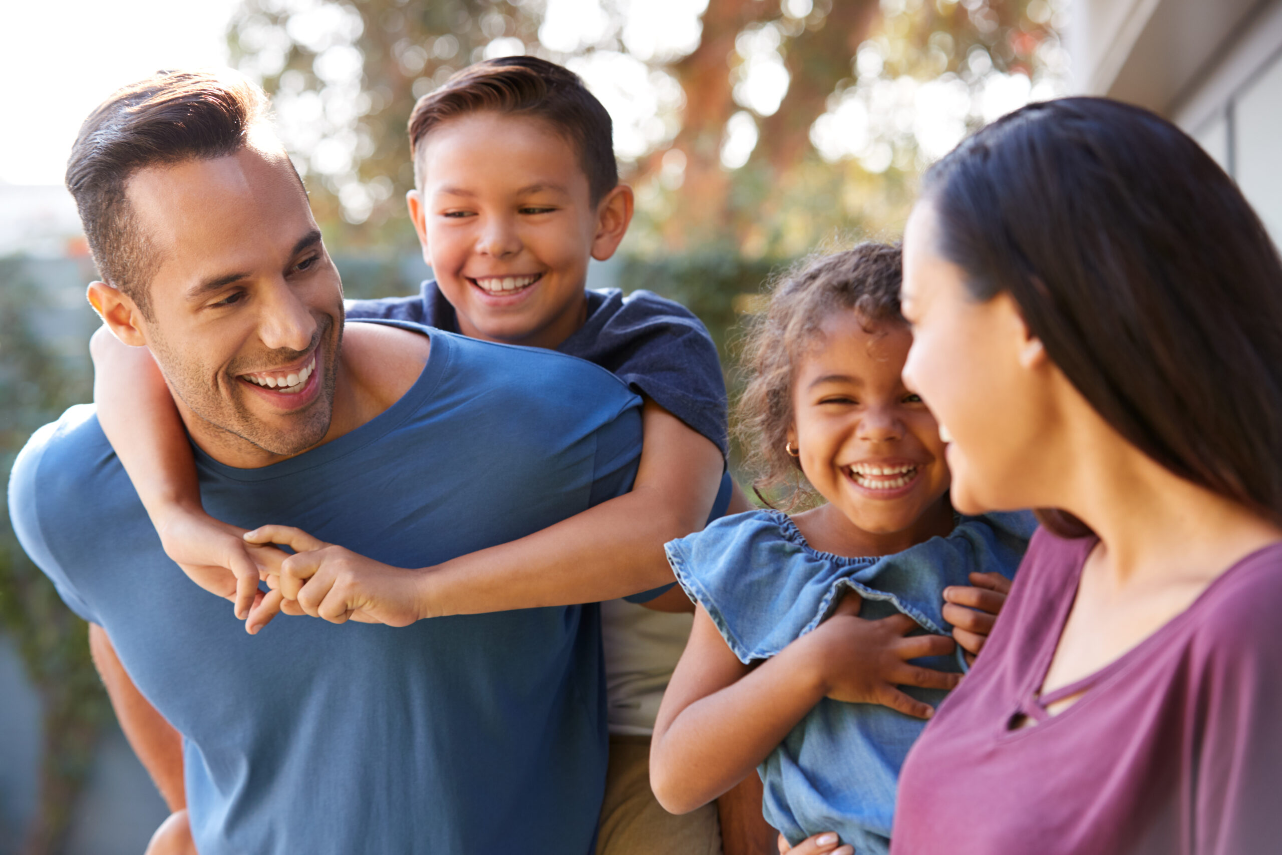 Smiling Hispanic Family With Parents Giving Children Piggyback Rides In Garden At Home Hispanic family smiling and giving their children piggyback rides in the garden at home